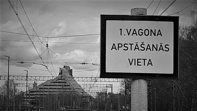 Dzelzceļa zīme. Balta plāksne ar uzrakstu melniem burtiem pirmā vagona apstāšanās vieta. Melnbalta fotogrāfija.