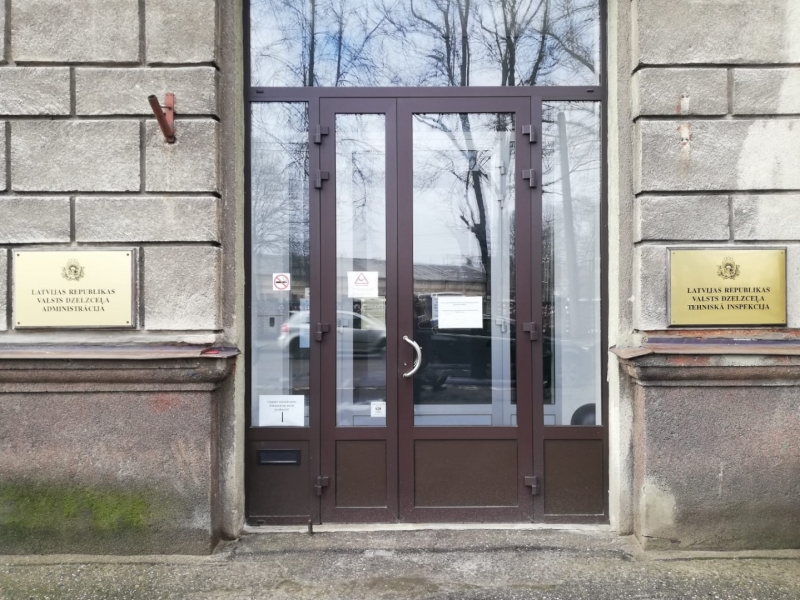 Attēls, kurā ir fragments no mājas fasādes, centrā ir iestādes brūnās krāsas durvis ar lieliem stikliem, kreisajā pusē centrā uz zelta krāsas plāksnes augšpusē attēlots “Latvijas mazais papildu ģerbonis”, bet zem tā melnā krāsā uzraksts “Latvijas republikas Valsts dzelzceļa Administrācija”, bet labajā pusē centrā uz zelta krāsas plāksnes augšpusē attēlots “Latvijas mazais papildu ģerbonis”, bet zem tā melnā krāsā uzraksts “Latvijas republikas Valsts dzelzceļa tehniskā inspekcija”.  