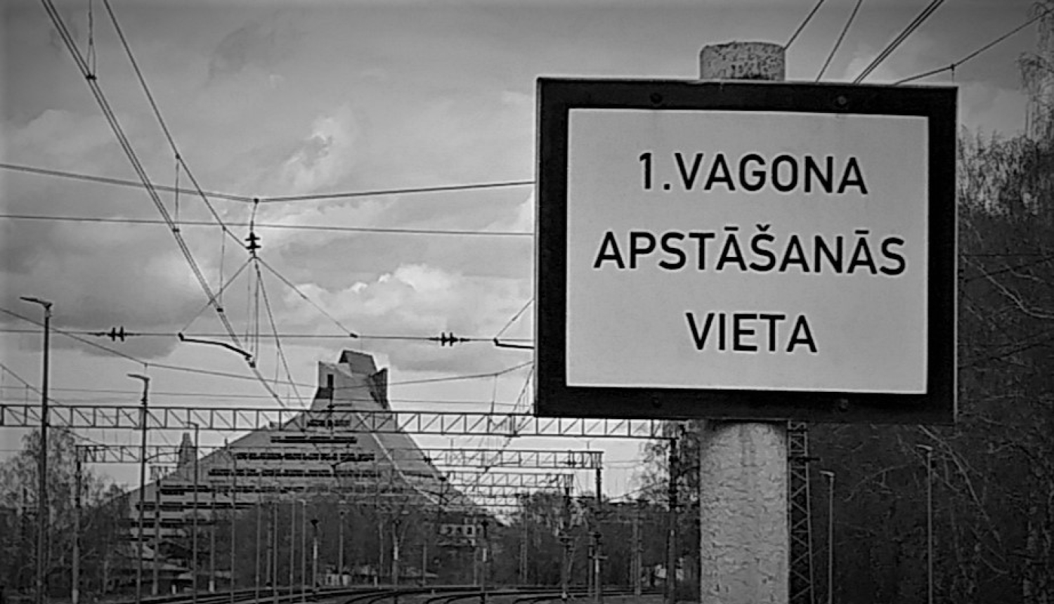 Dzelzceļa zīme. Balta plāksne ar uzrakstu melniem burtiem pirmā vagona apstāšanās vieta. Melnbalta fotogrāfija.